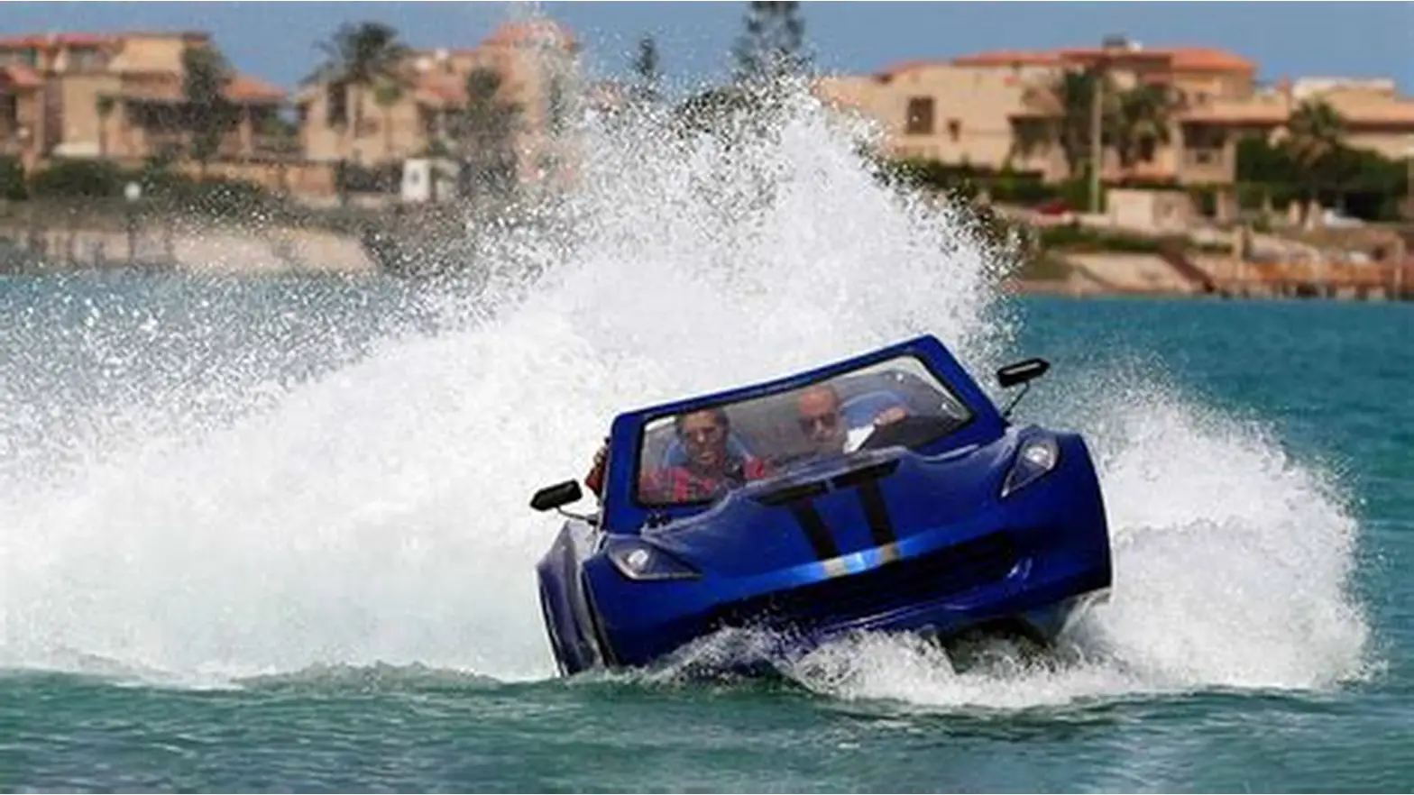 Blue car-shaped boat speeding through water, creating a massive white splash near a shoreline.