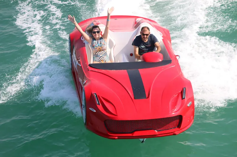 A red sports car-shaped boat speeds through turquoise water with two happy passengers inside.